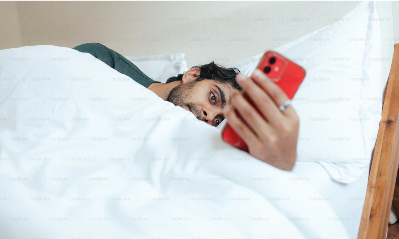 Un homme dans son lit qui regarde son téléphone au-dessus des couvertes.