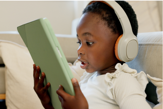 Un enfant de 2-4 qui avec des écouteurs qui regarde une tablette assez proche de son visage assis sur un sofa blanc.