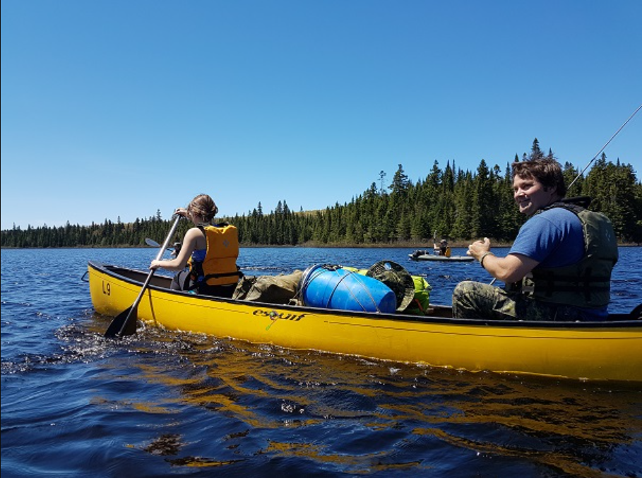 Un canot jaune rempli de sacs sur un lac opéré par un homme et une femme. Il fait soleil et on voit la forêt à l'horizon.