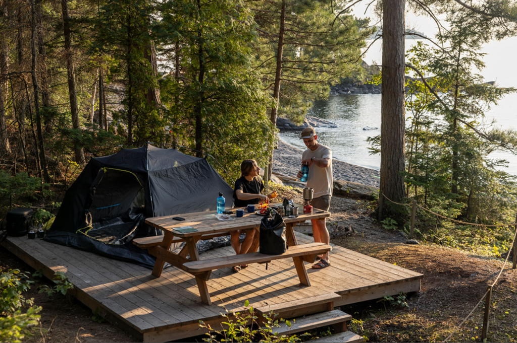 Deux hommes qui préparent leur déjeuner après une nuit de camping au parc national du mont tremblanc. Ils sont sur une table en bois classique à côté de leur tente noire dans la forêt sur le bord de l'eau.