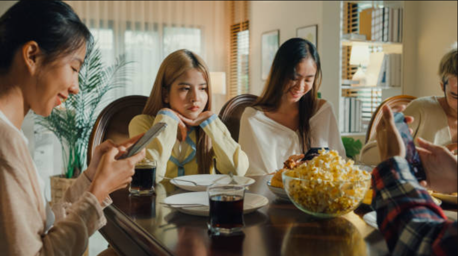 Une famille à table pour souper. Toute la famille est sur son téléphone, sauf une femme qui regarde dans le vide.