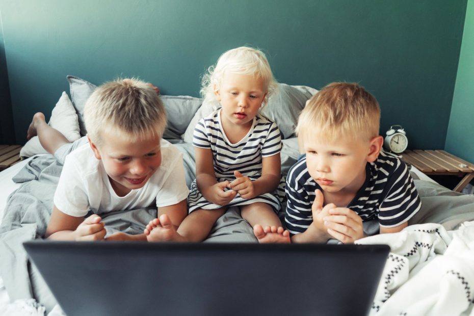 Trois enfants blonds très petits couchés sur un lit blanc qui regardent tous une télévision au centre des trois enfants.