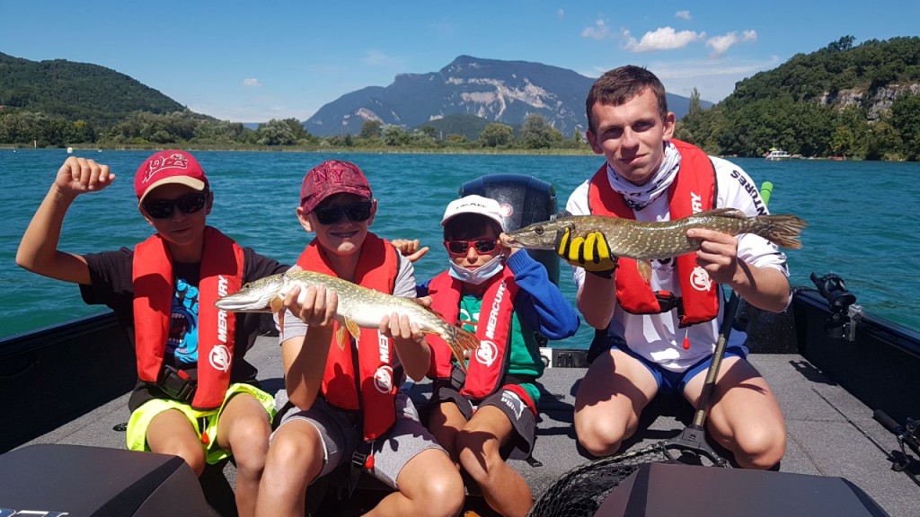 Un groupe de 4 adolescents en bateau qui montrent ce qu'ils ont attrapé pendant la pêche. Deux d'entre eux ont des poissons, le lac et le paysage sont visibles à l'horizon. C'est magnifique!