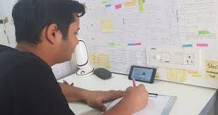un étudiant qui écrit dans un cahier tout en regardant un vidéo sur son téléphone collé au mur rempli de feuilles d'études.